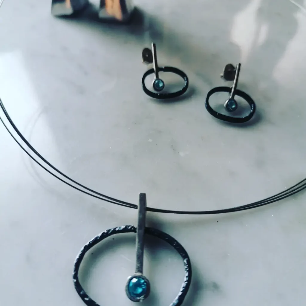 Un conjunt de joies contemporani amb un collaret i arracades a joc, ambdues peces presenten un disseny abstracte que combina un cercle o oval amb una barra i una petita pedra blava. Les joies tenen un acabat fosc i mat, amb la pedra blava que destaca com a punt focal. El conjunt està col·locat sobre una superfície de marbre blanc, amb un objecte metàl·lic reflectant al fons.
